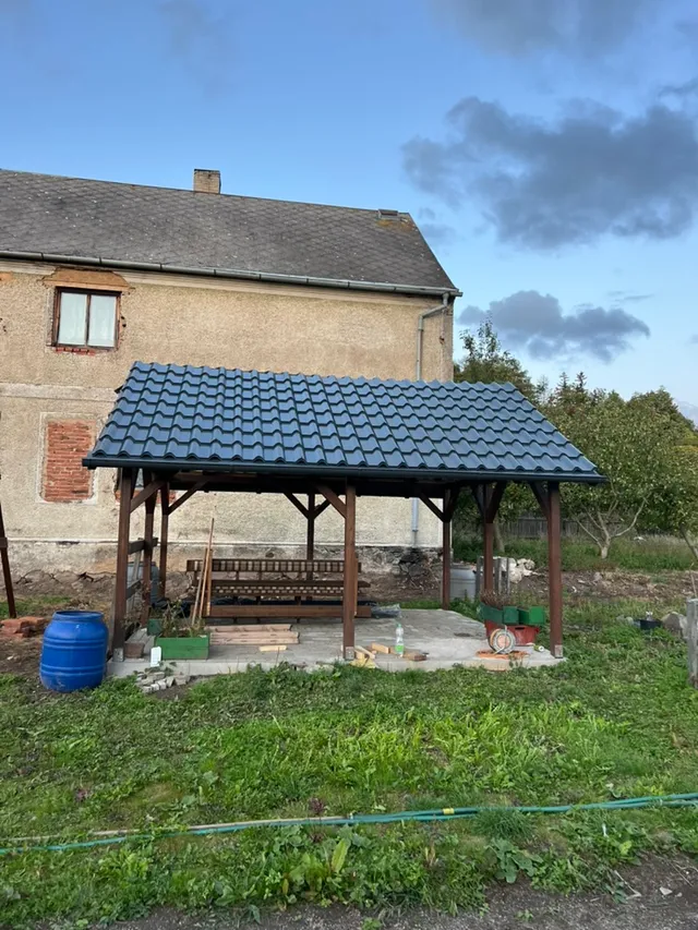 Hotová pergola u sousedního domu — sedlová střecha v taškovém profilu, pod ní dlážděná podlaha se vzorem. Konečně místo, kde trávit čas.