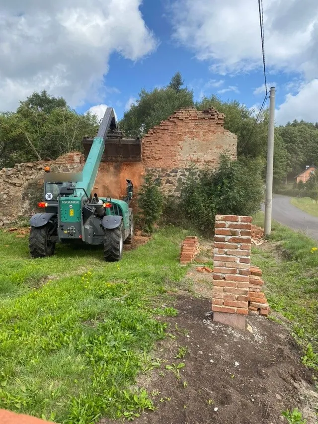 Teleskopický manipulátor Kramer boří štítovou stěnu ruiny. Vpravo už stojí první cihlový sloupek budoucího plotu.