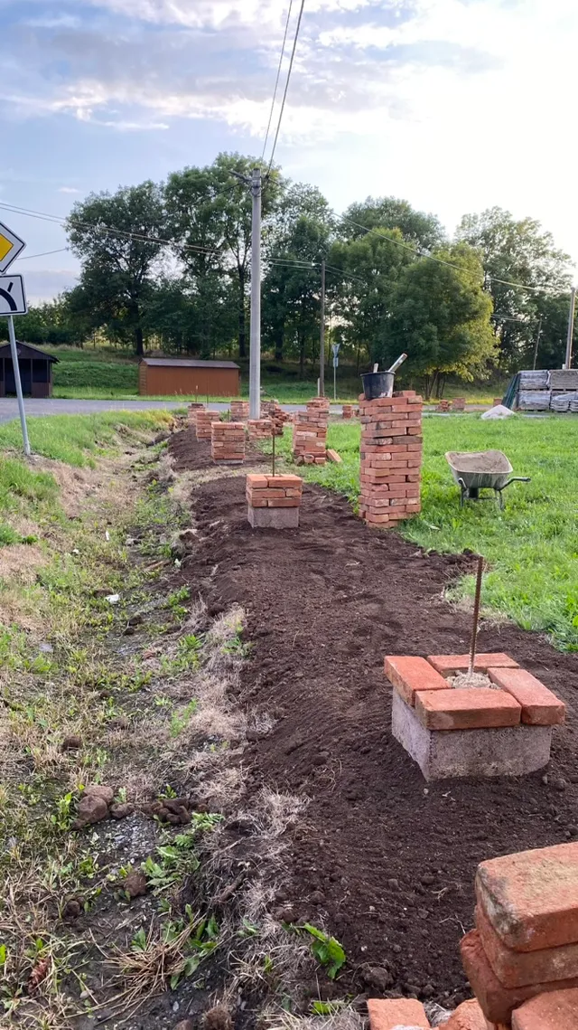 Cihlové sloupky ve výstavbě — armování, kolečko s maltou a řada budoucích patek podél příkopu.