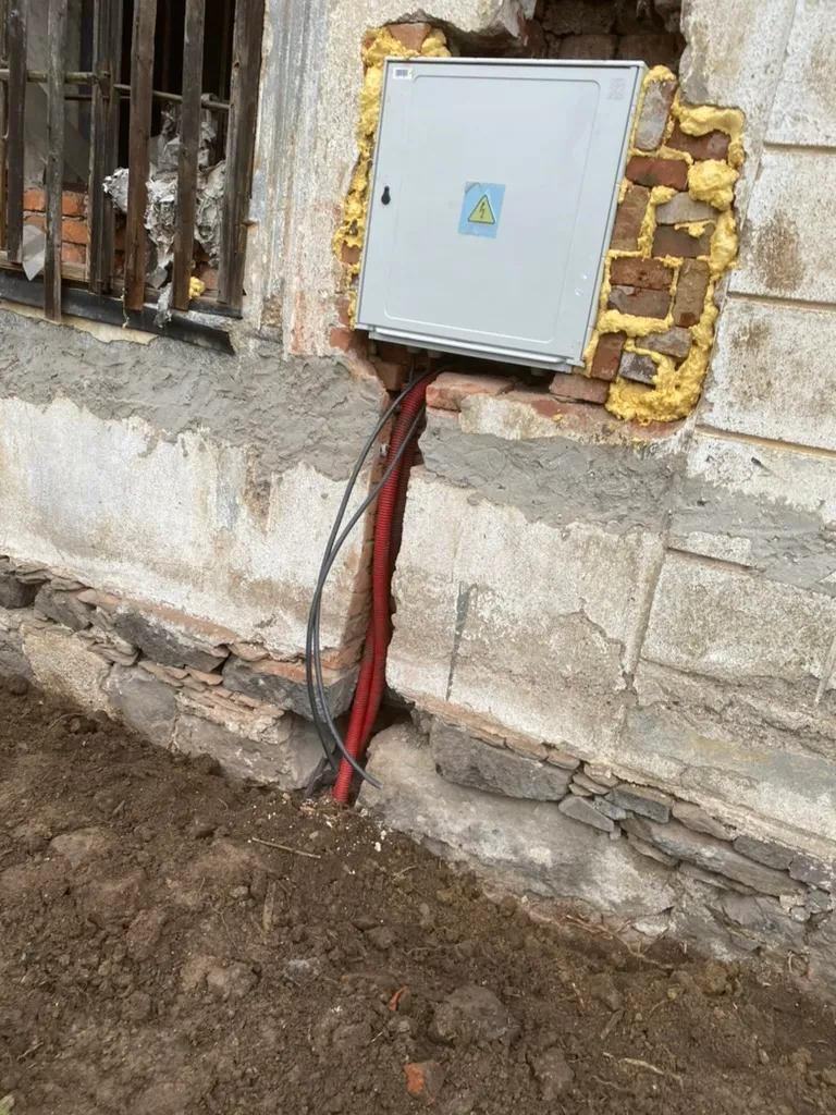 Elektroměrový rozvaděč zvenku, kabel v červené chráničce dolů do výkopu. Oficiální přípojka.