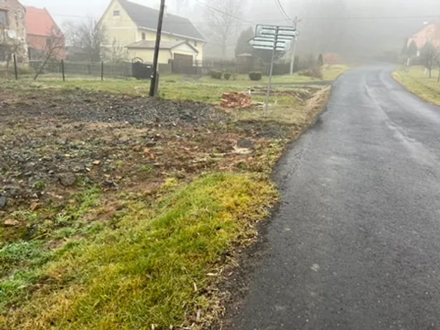 Uklizený pozemek podél cesty. Po ruině zbyla jen tmavá plocha s drobnou sutí — a spousta prostoru.